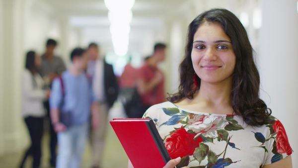 Portrait of a happy female indian student standing in a busy hallway ...