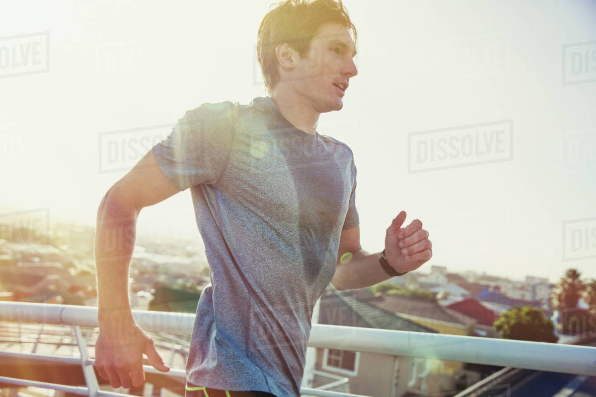 Sweaty male runner running on sunny urban footbridge at sunrise Stock
