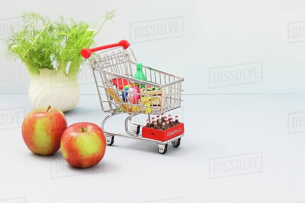 A mini shopping trolley filled with toy foodstuffs next to apples and a