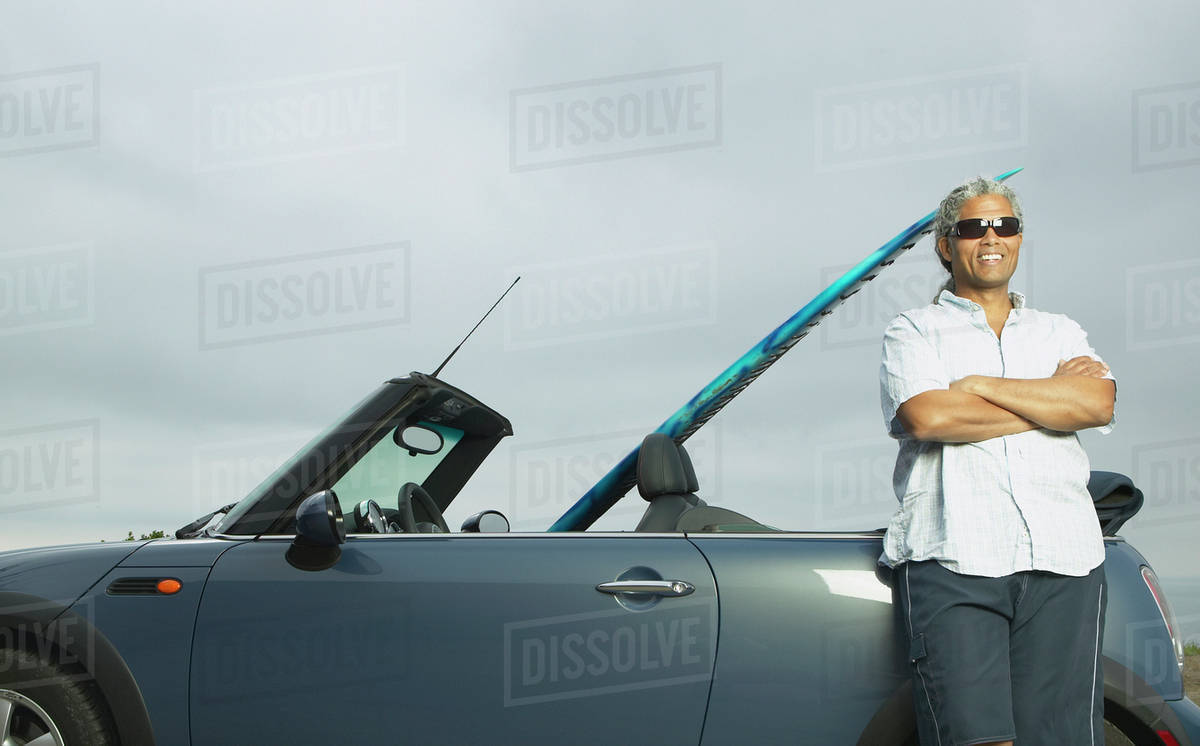 Senior man leaning on a convertible with a surfboard in it Stock