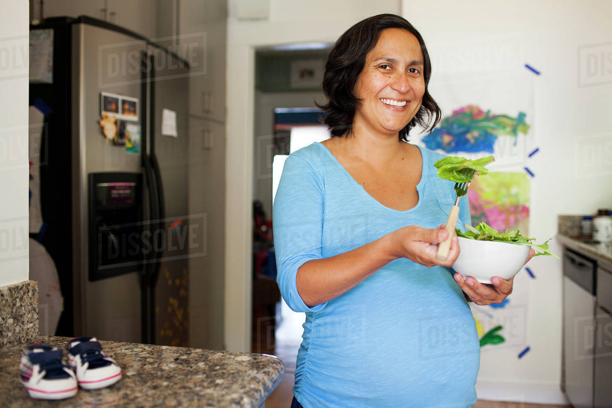 Pregnant Hispanic woman eating salad in kitchen Stock Photo Dissolve