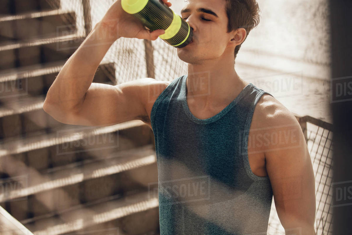 Shot of muscular young man drinking water by the beach after workout