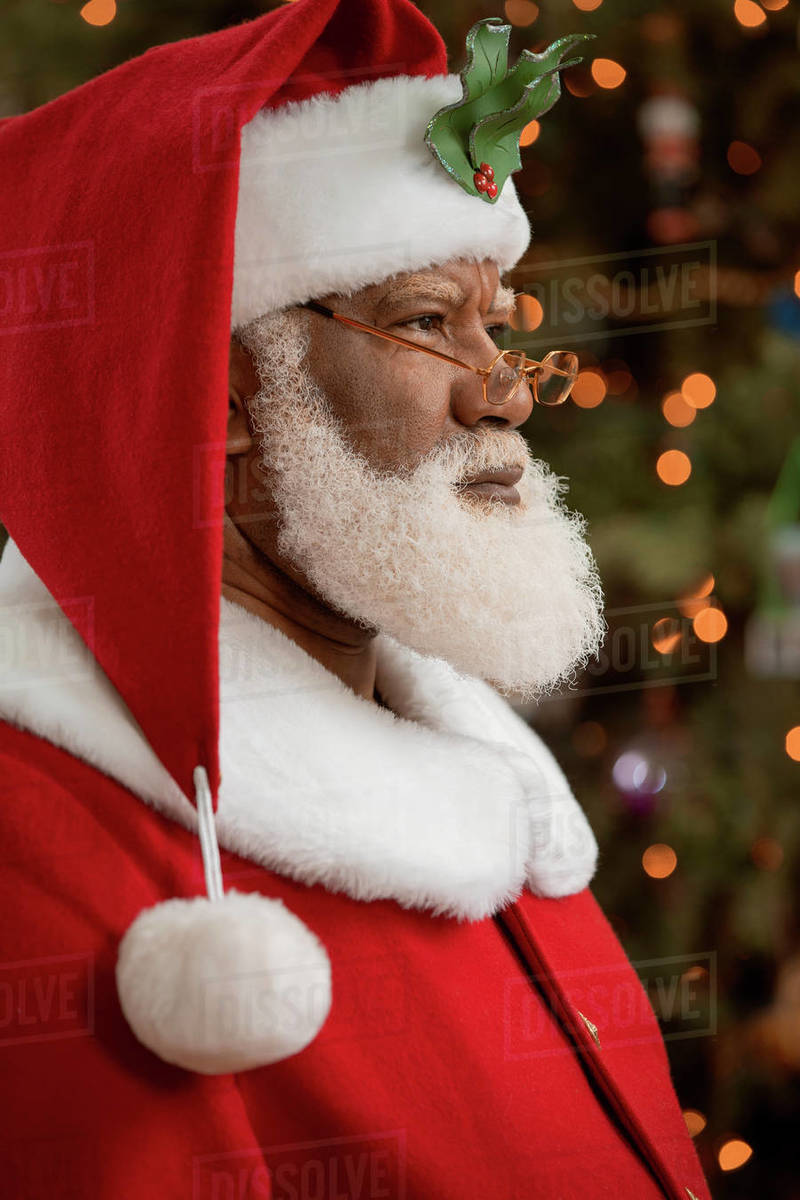 An African American man dressed as Santa Claus sitting in front of a Christmas tree turned profile, looking off camera. Royalty-free stock photo