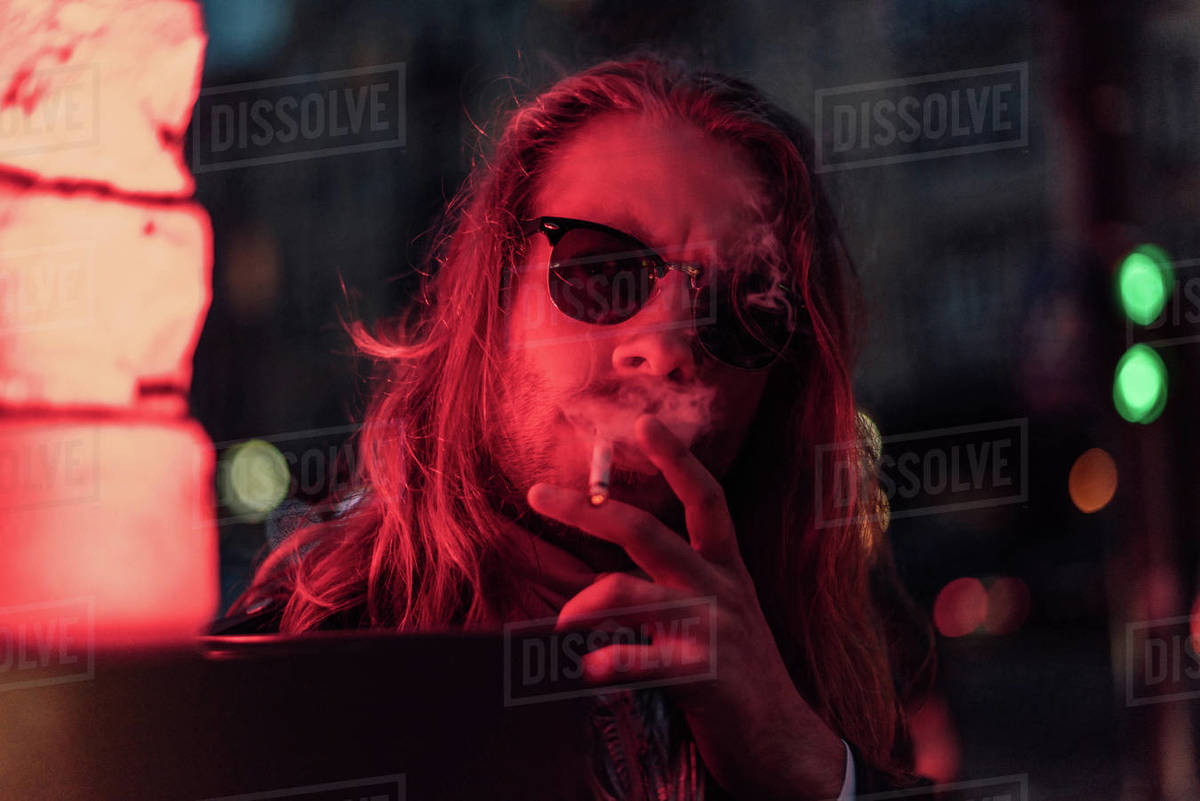 Handsome young man in sunglasses smoking cigarette under red light on