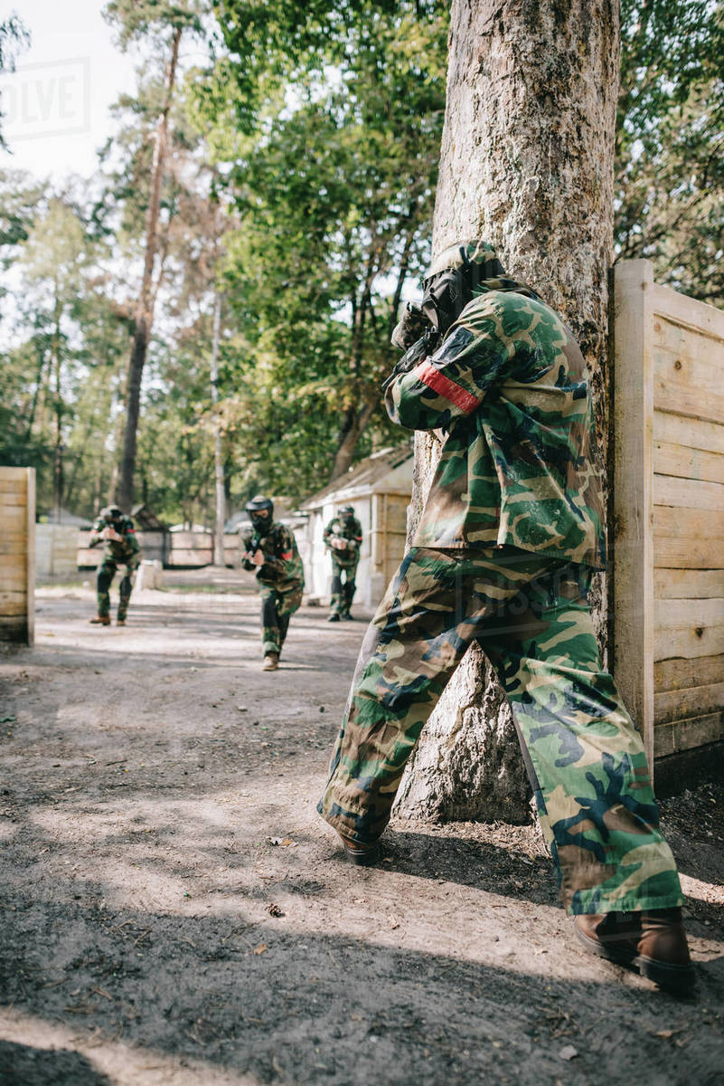 Rear view of paintball player in camouflage shooting by marker gun in