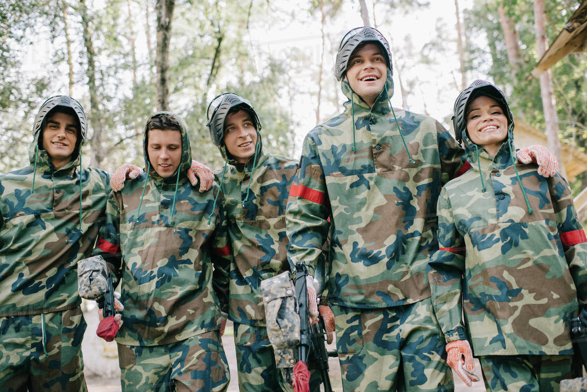 Happy paintball players in camouflage with marker guns embracing each other outdoors Stock