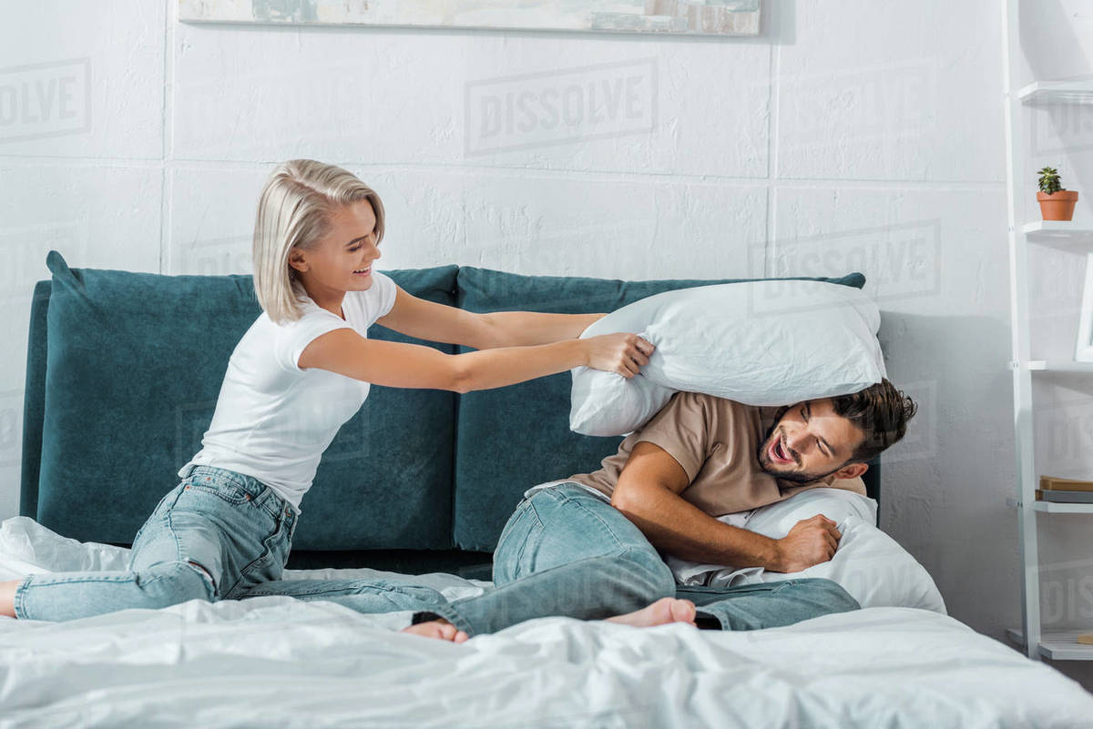 Happy Girlfriend Beating Boyfriend With Pillow On Bed In Bedroom