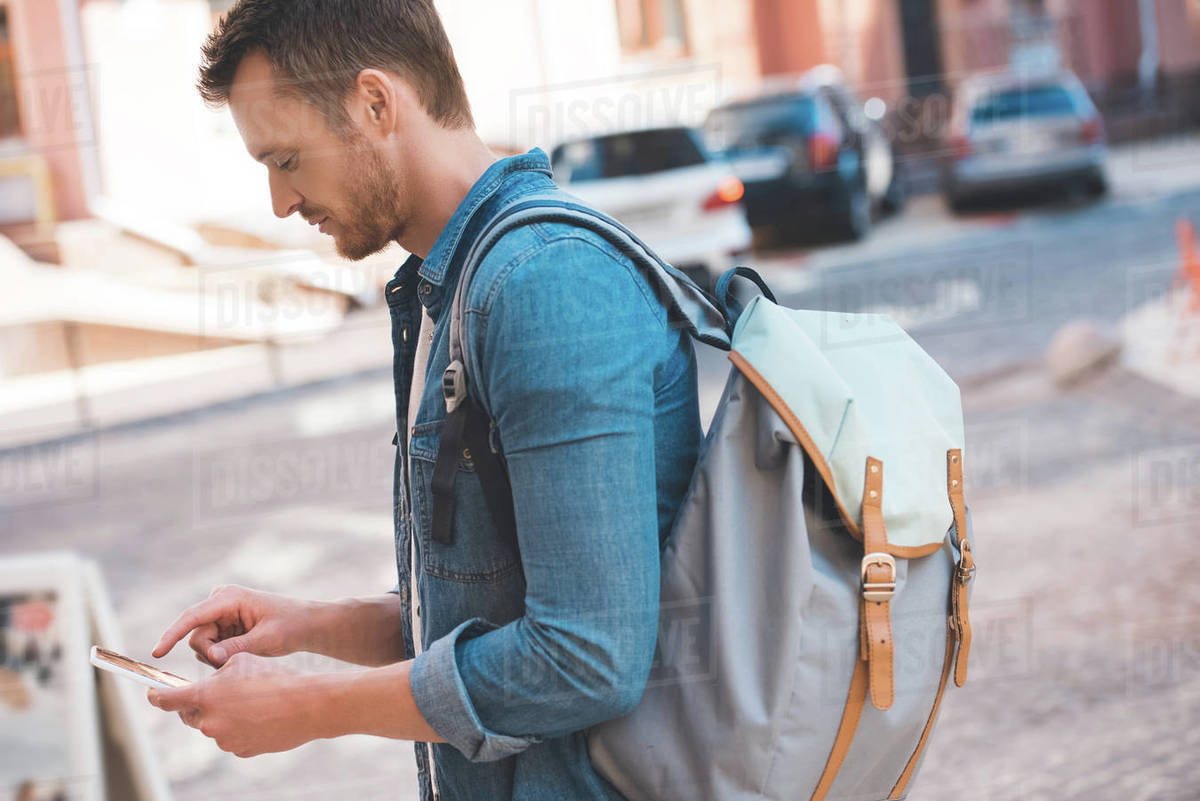 Handsome young man with backpack walking by street and using smartphone Stock Photo Dissolve