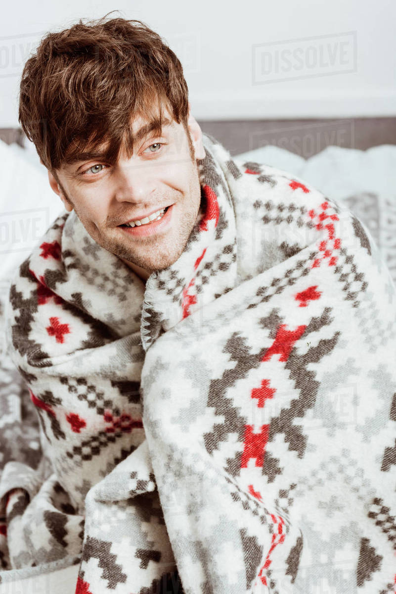 cheerful sick young man wrapped in blanket sitting on bed at home