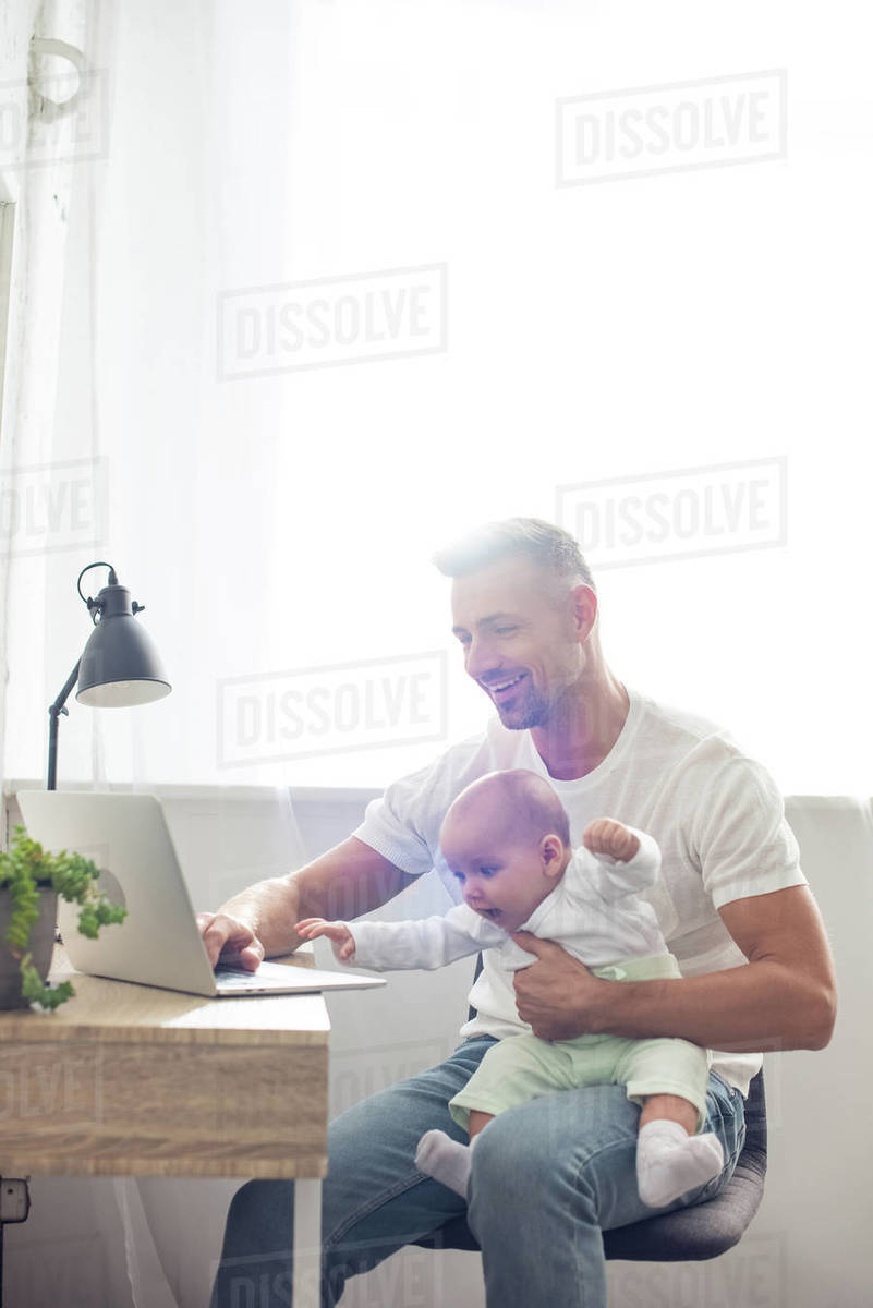 Happy father sitting on chair, holding baby daughter and using laptop