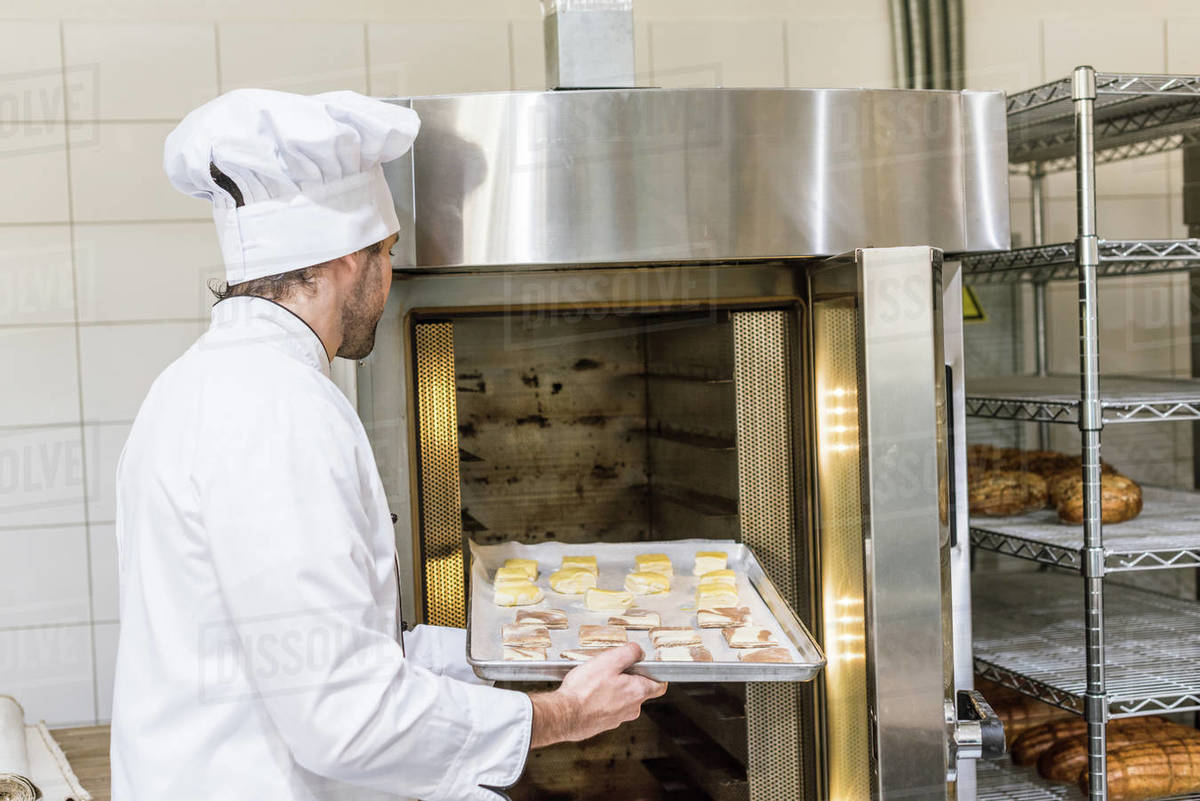 male baker in chefs uniform putting raw dough in oven Stock Photo