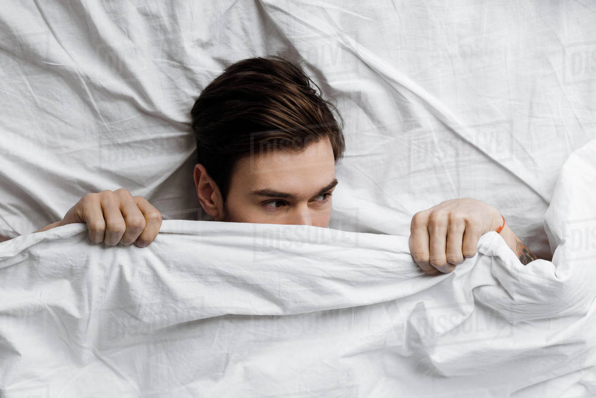 top view of handsome young man hiding under blanket in bed at home and