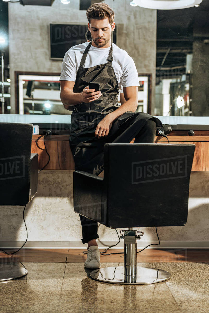 Handsome Young Hairstylist In Apron Using Smartphone In Beauty