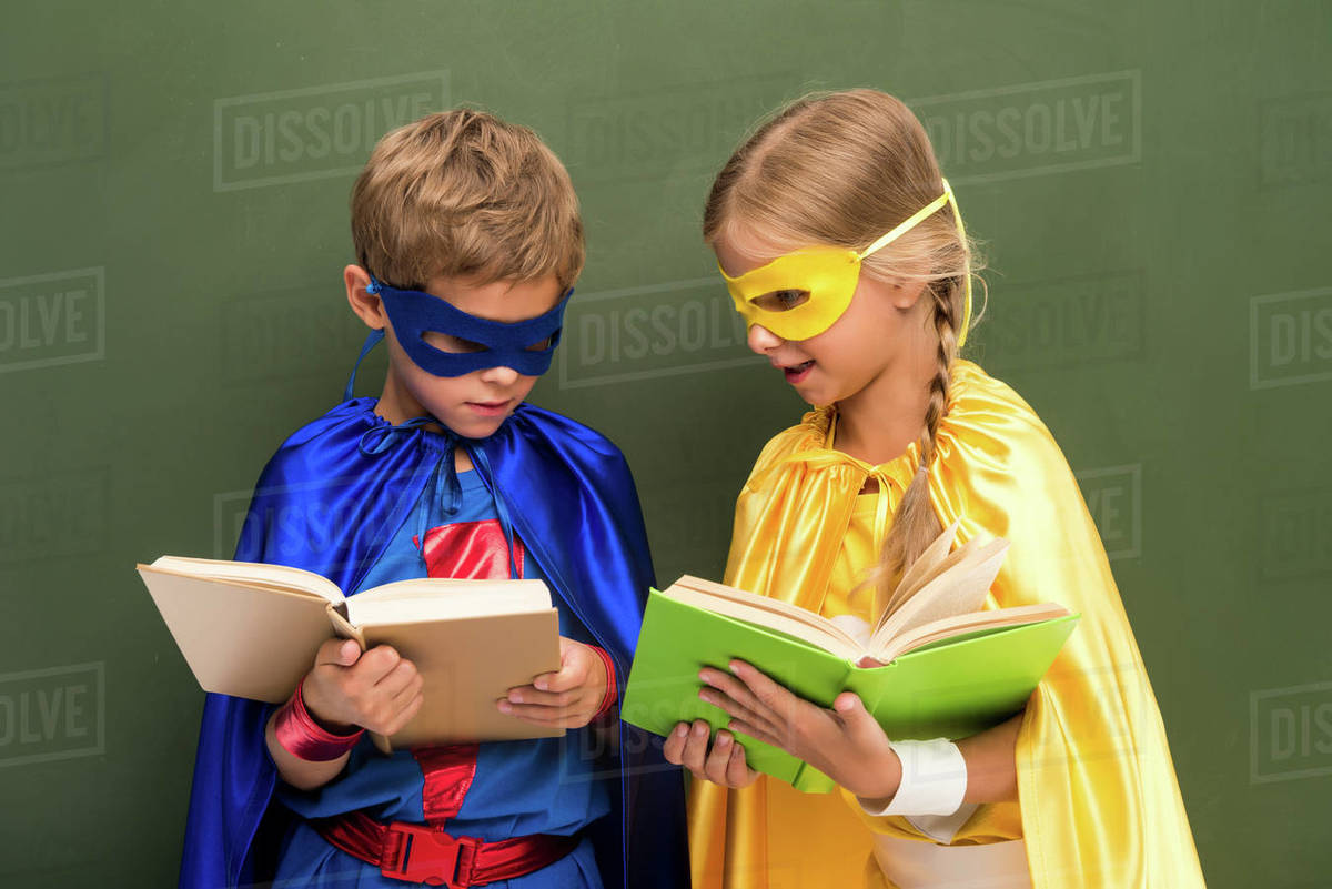 Little pupils in superhero costumes reading books, chalkboard behind