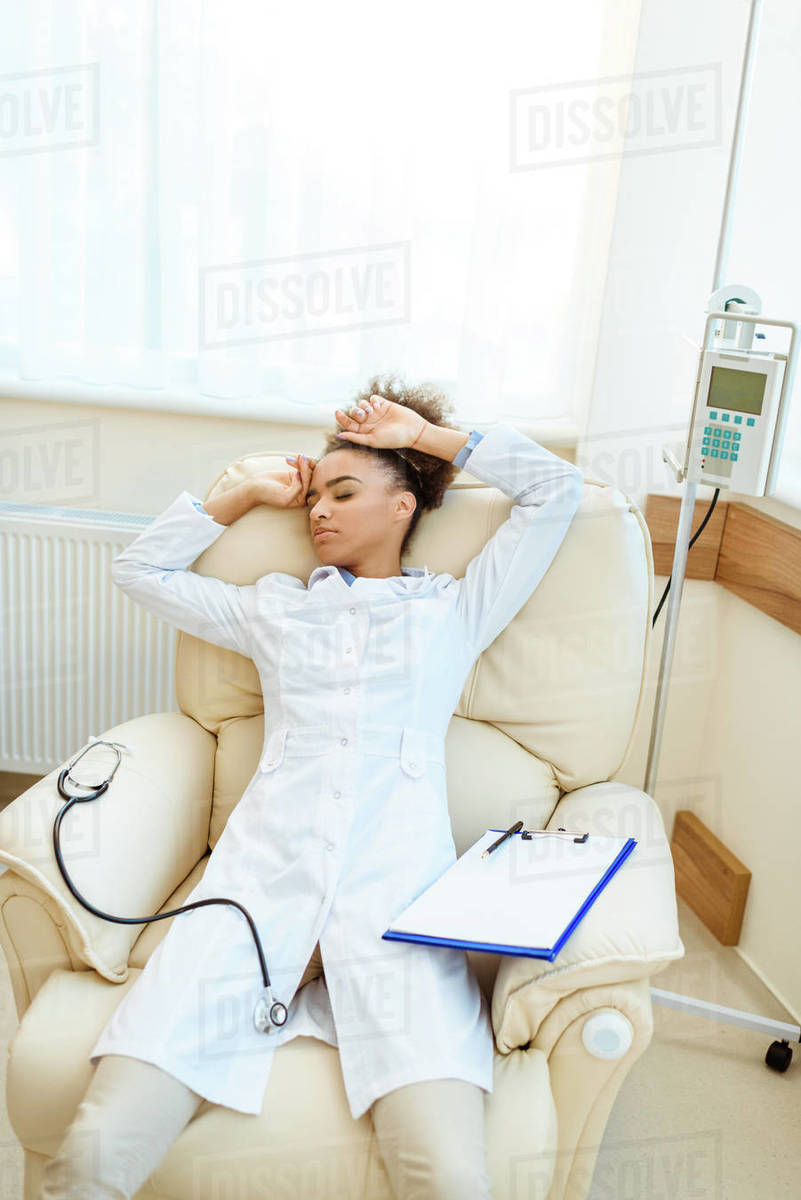 Young African American doctor lying in armchair exhausted with stethoscope and clipboard Stock