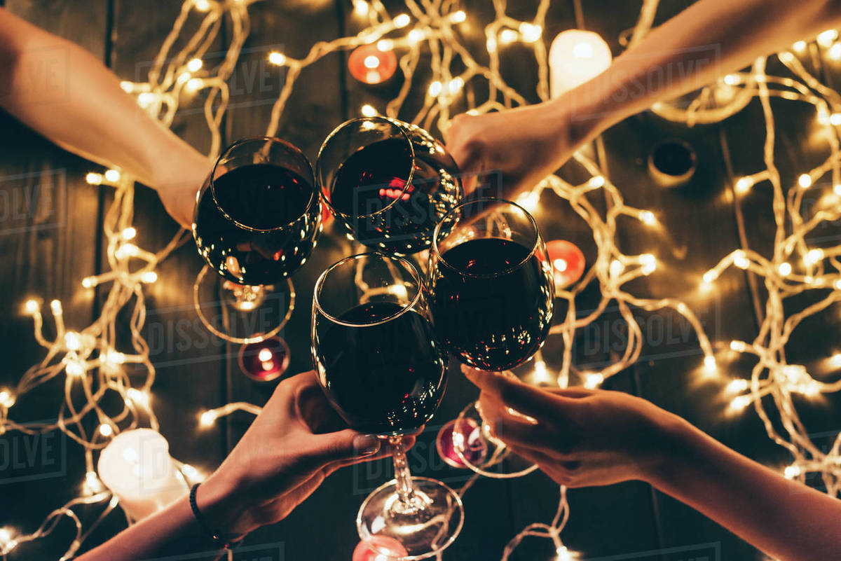 Cropped shot of four people clinking glasses with red wine over wooden  table with fairylights decorations - Royalty-free Stock Photo | Dissolve