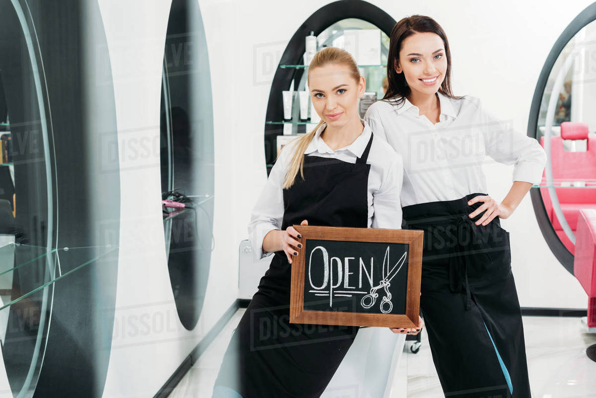 Two beautiful hairdressers standing with sign open Stock Photo Dissolve