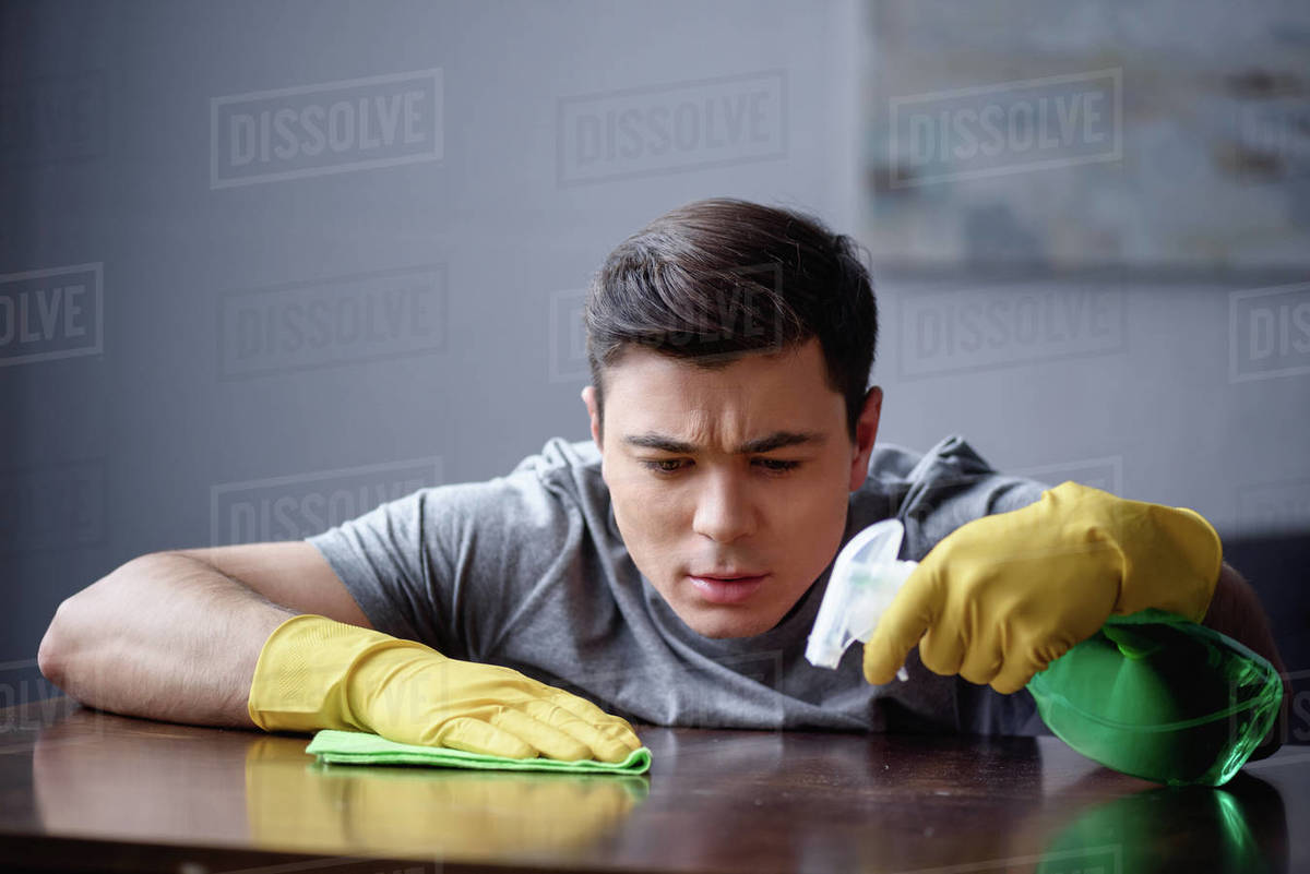 Handsome man dusting table in living room with spray bottle and rag