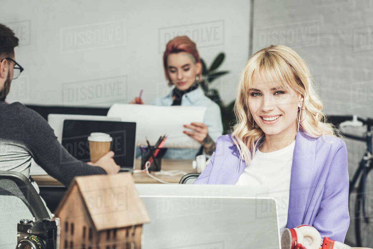 Selective focus of smiling blogger at workplace with colleagues behind ...