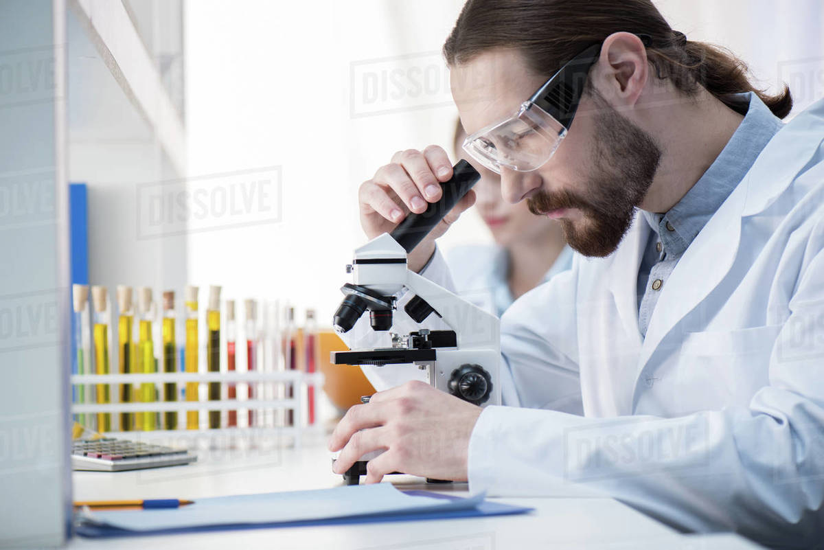 Young man chemist in protective glasses looking in microscope in lab