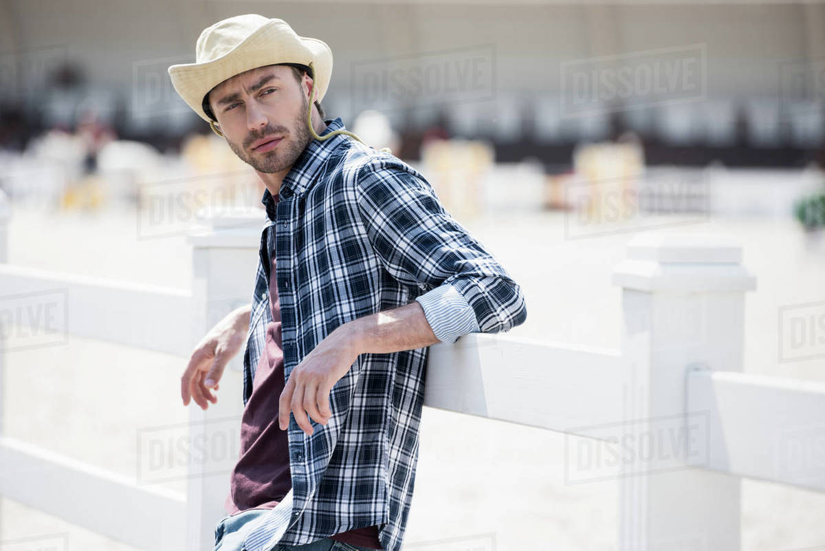 Handsome young man in cowboy hat leaning at wooden fence and