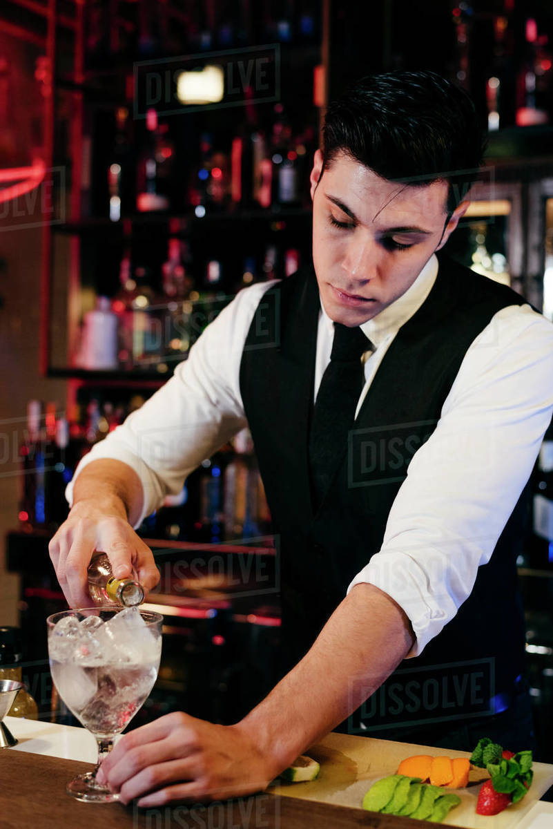 Young elegant barman working behind a bar counter mixing drinks with