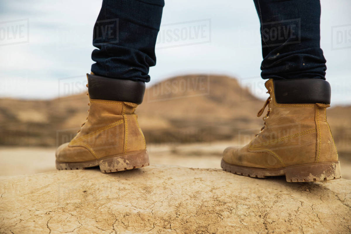 Legs on faceless traveler in brown boots and blue jeans standing on dirty sandy road with
