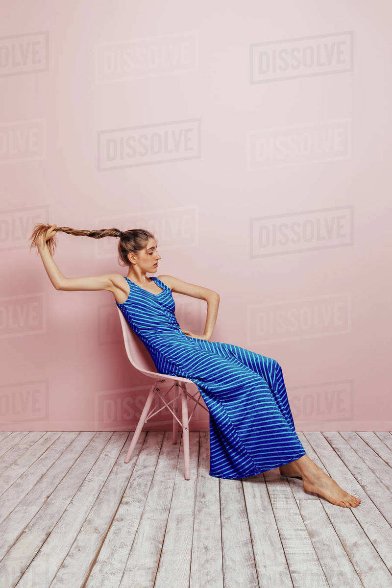 Side view of young woman in blue dress barefoot looking at camera holding  ponytail while sitting in chair and looking away on minimalist pink  background - Royalty-free Stock Photo | Dissolve