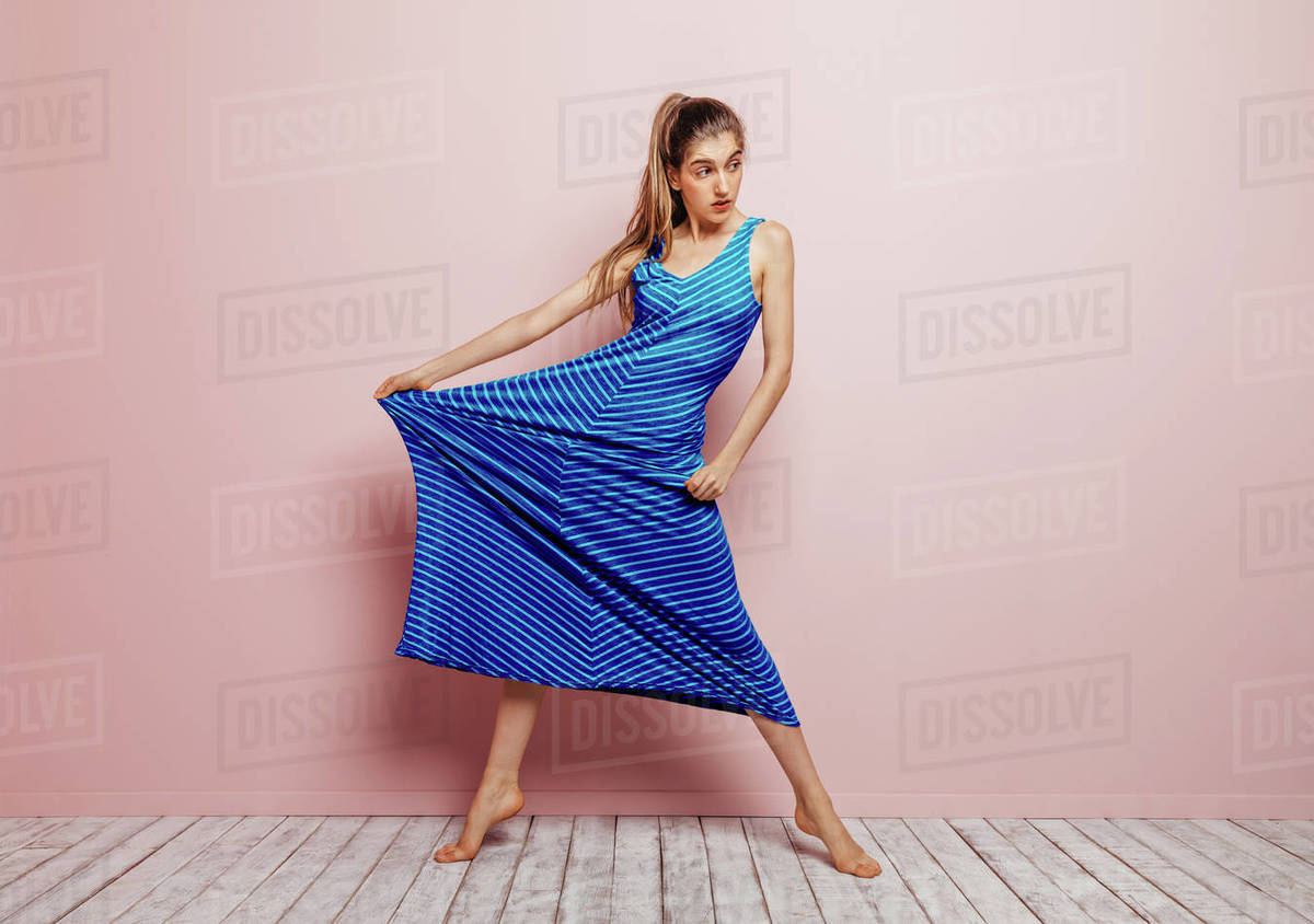Young woman barefoot looking away stretching side of a blue dress while  standing on minimalist pink background - Royalty-free Stock Photo | Dissolve