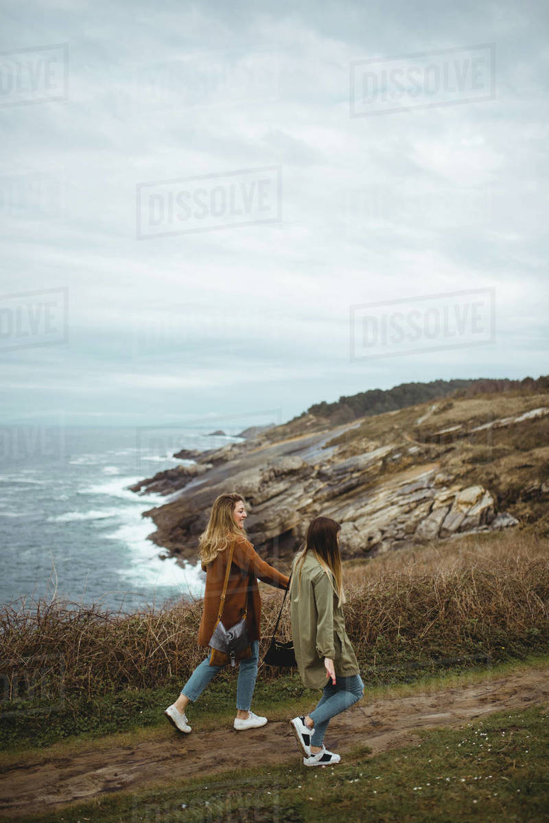 Cheerful young women holding hands and laughing while walking on narrow ...