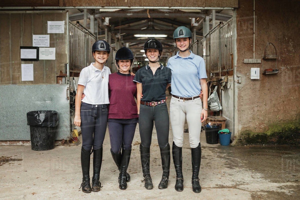 Group of friendly female equestrians in uniform hugging near barn in