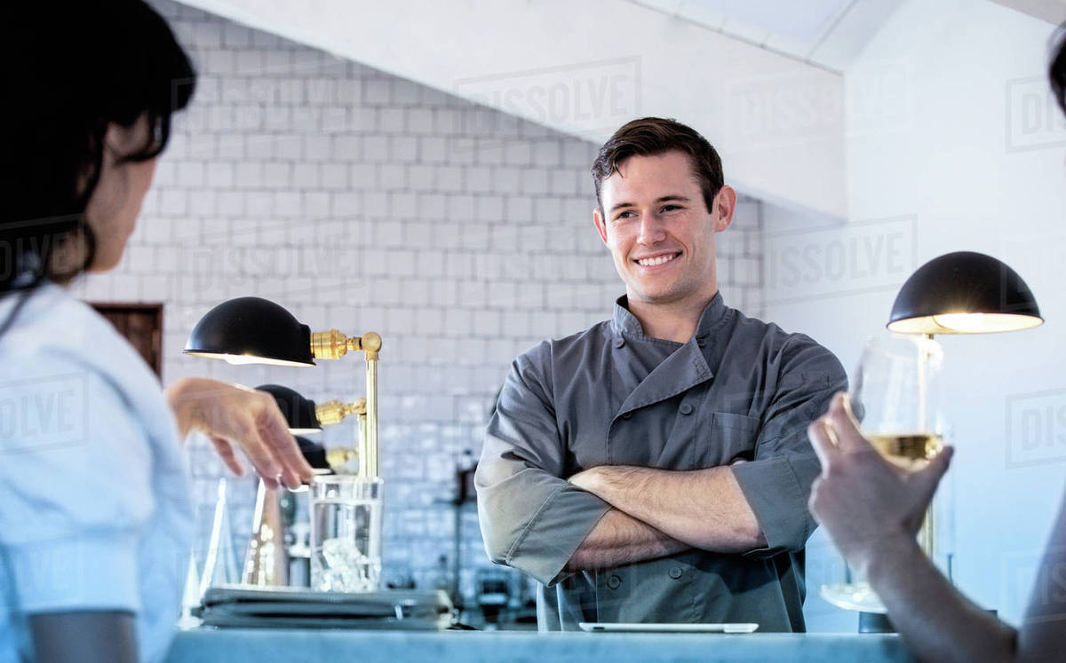 Barman standing behind bar, two customers sitting at bar with drinks