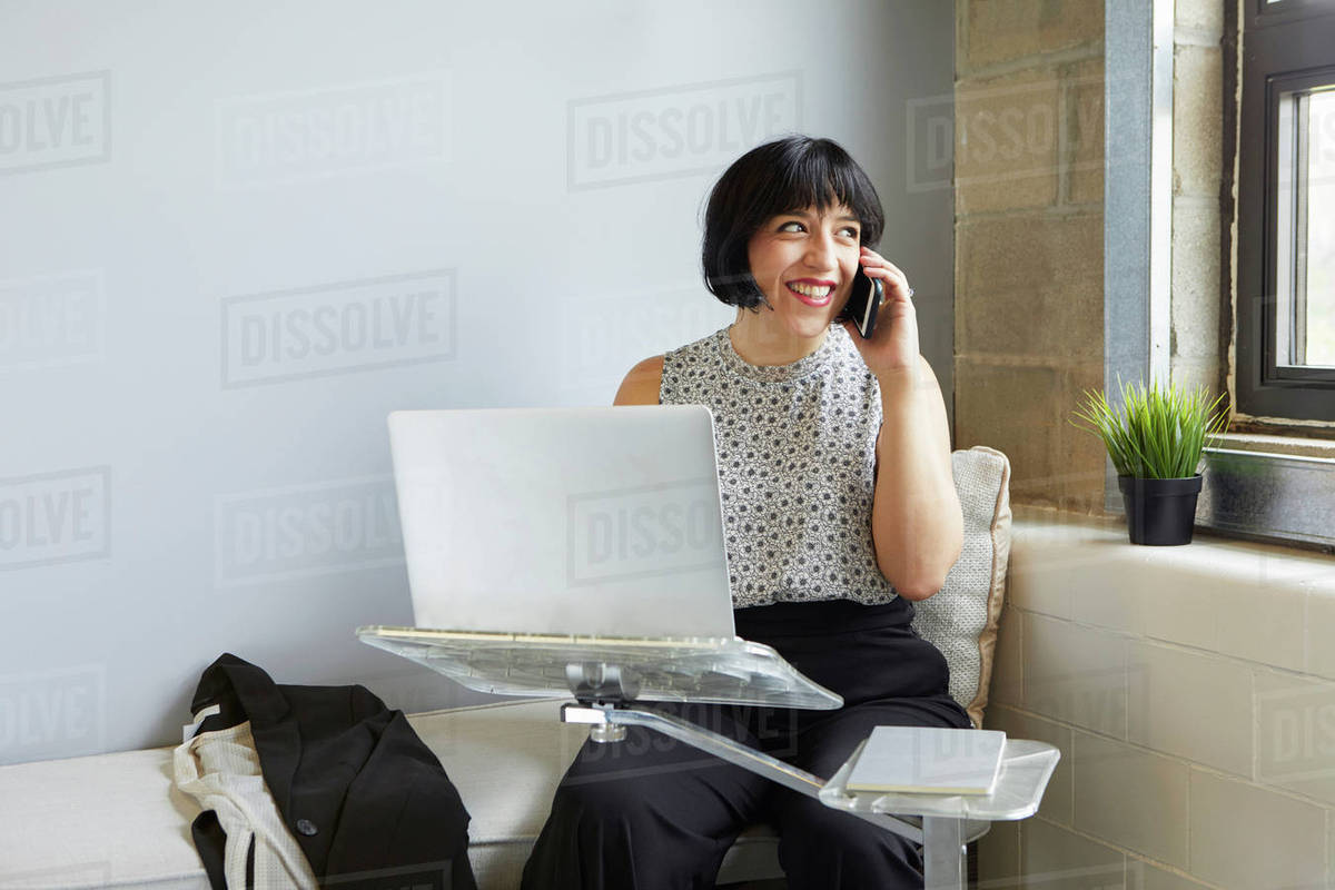 Woman sitting using laptop on laptop stand, speaking on smartphone
