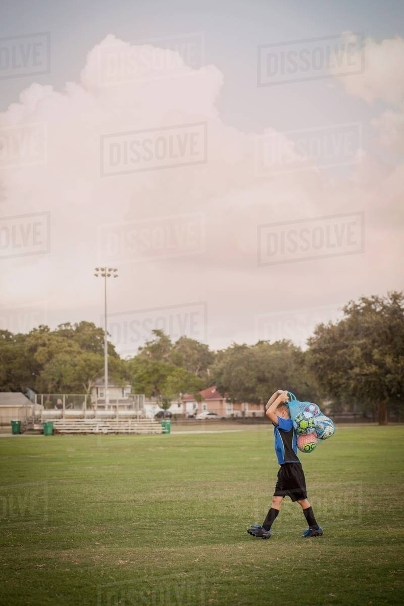 Boy football player carrying bag of footballs on practice pitch Stock Photo Dissolve