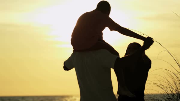 silhouette of happy african american family playing on beach vacation with son riding fathers shoulders at