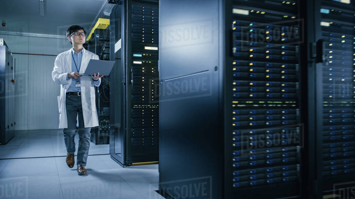 In Data Center Male IT Technician Wearing White Coat Walking alongside Server Racks, Uses
