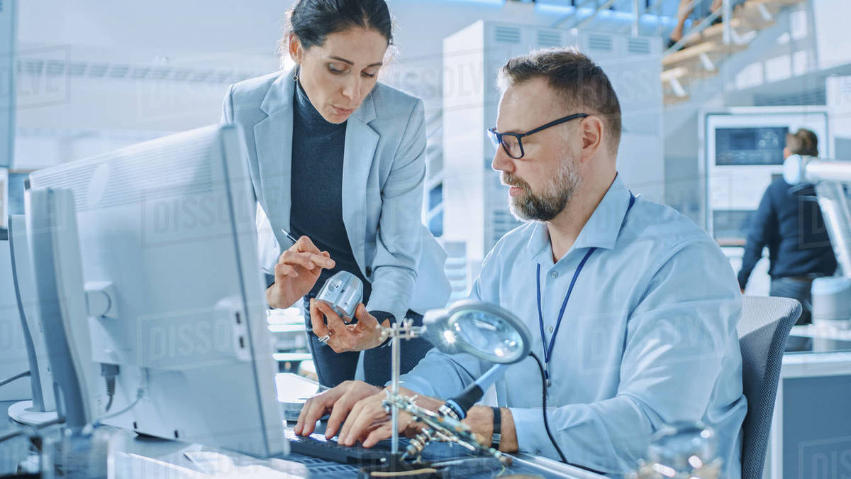 Female Industrial Engineer Talks with Electronics Specialist, She Holds Metal Component