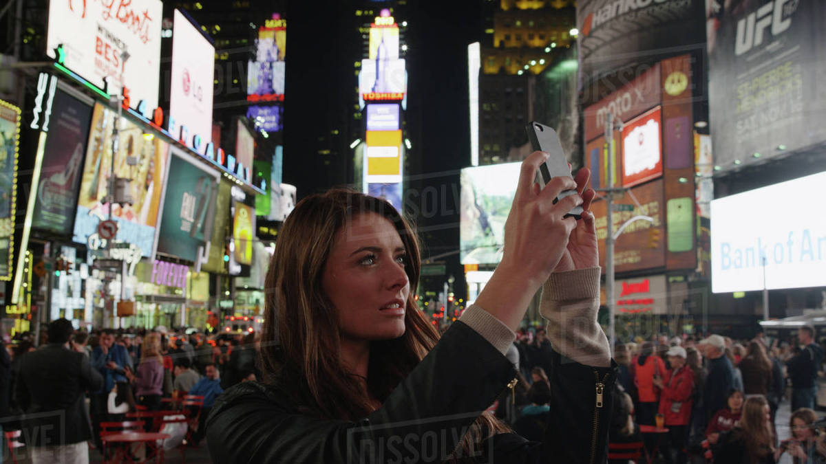 Tourist photographing iconic glowing billboard advertisements with cell phone in city intersection at night Royalty-free stock photo