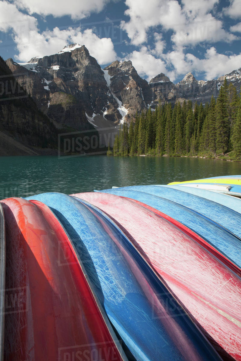 Row Of Canoes On Their Sides With A Lake Mountains Clouds And Blue Sky