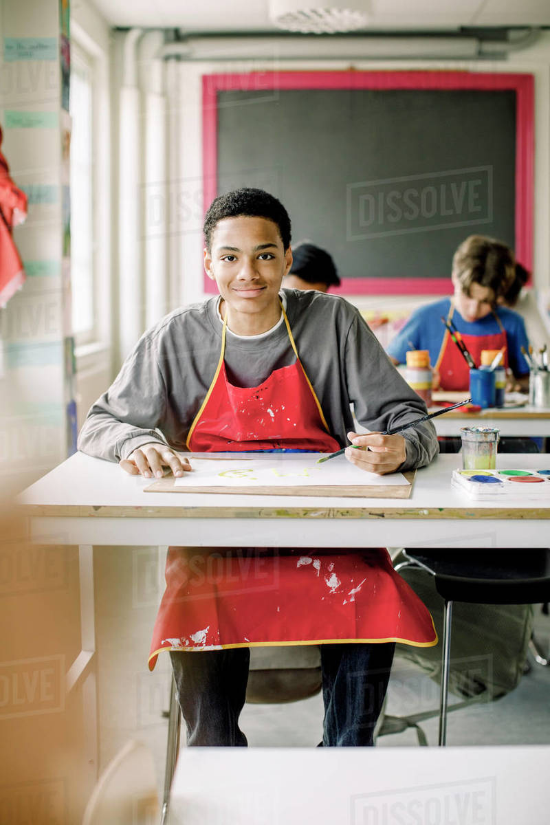 Portrait of smiling male teenage student sitting at desk during art