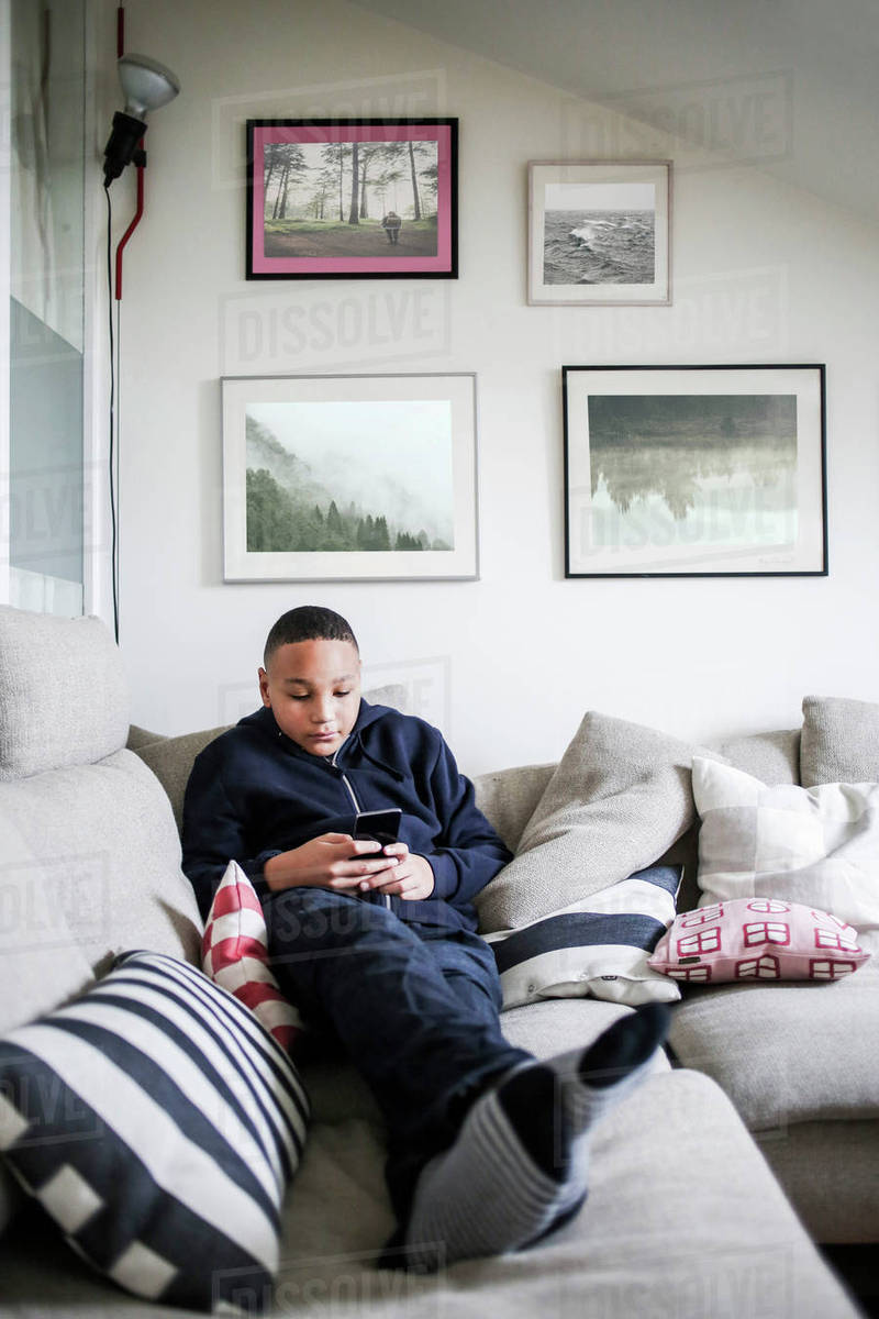 Full Length Of Age Boy Using Mobile Phone On Sofa At Home Stock Photo