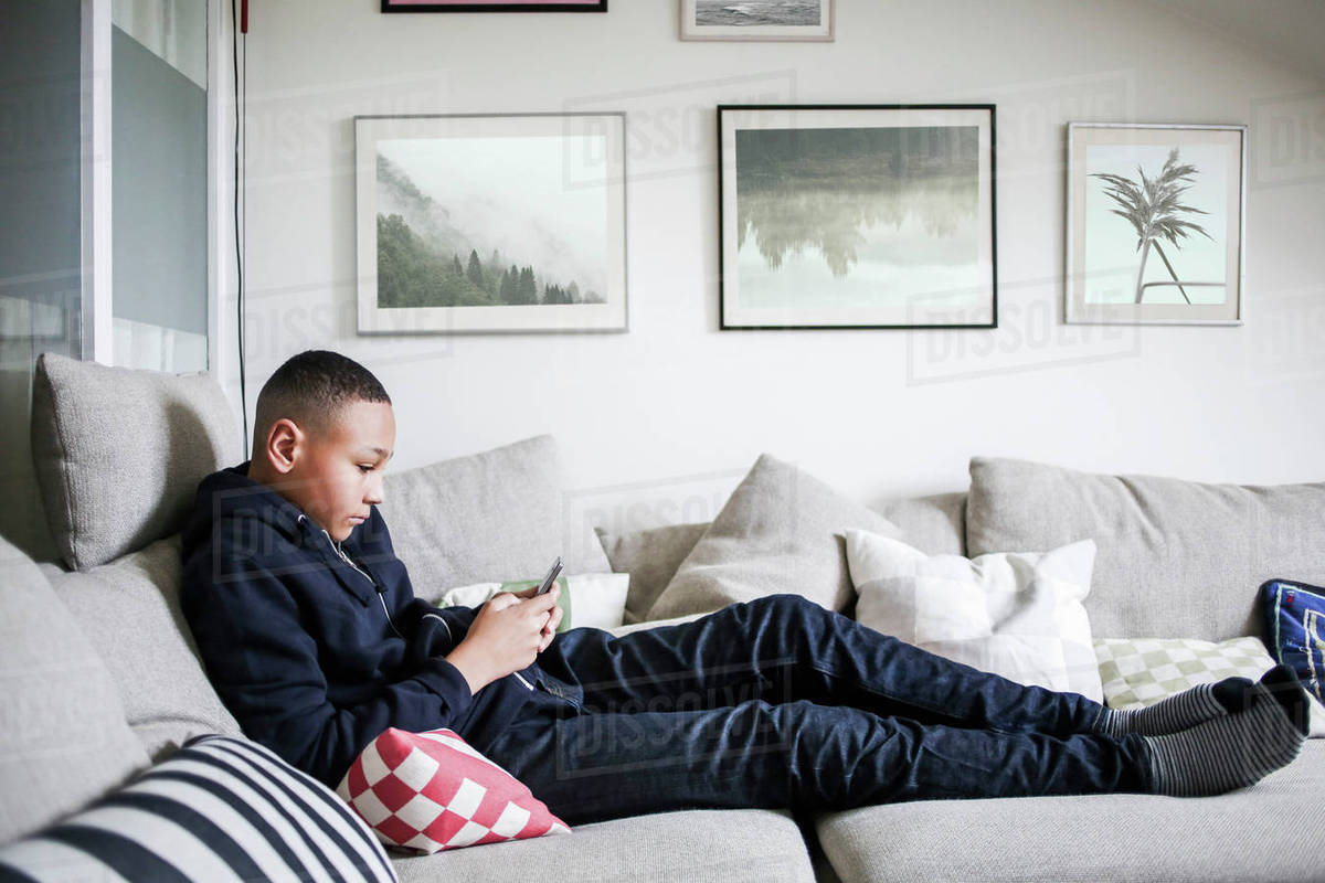 Full Length Side View Of Age Boy Using Mobile Phone On Sofa At Home Stock Photo