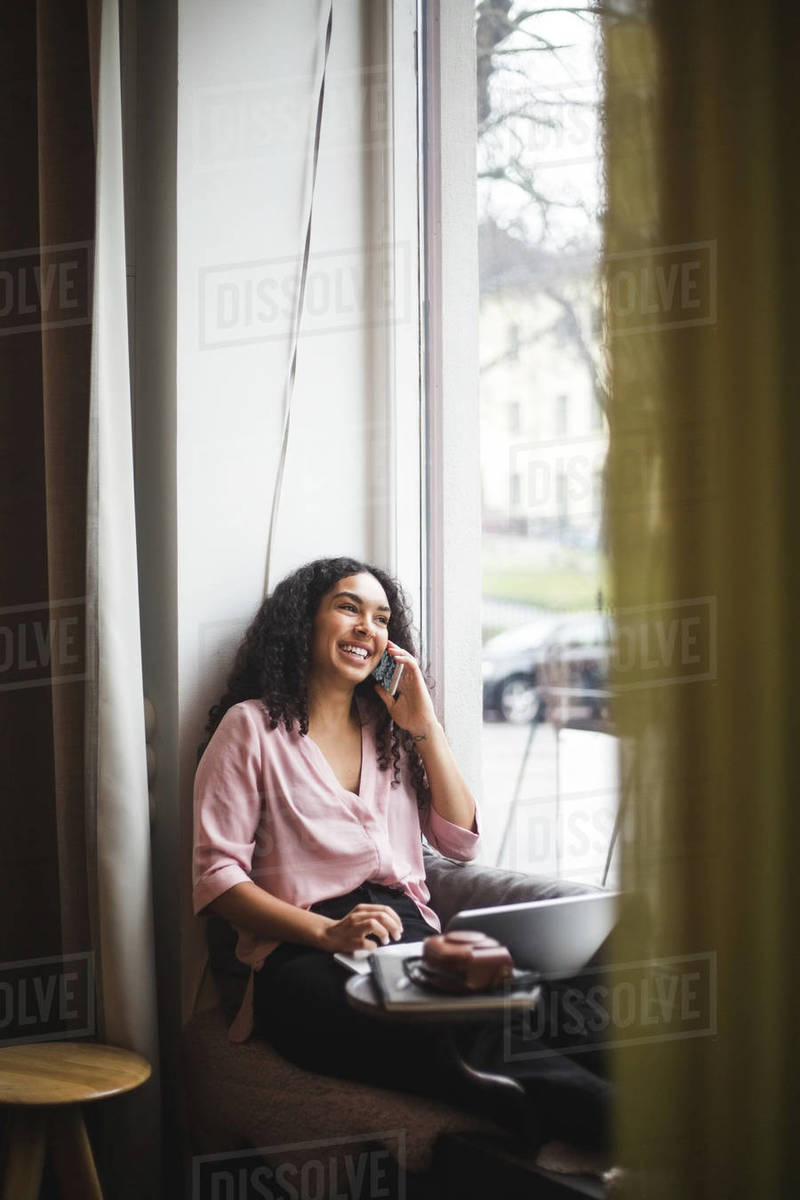 Smiling young female professional talking through smart phone sitting on window sill in office Royalty-free stock photo