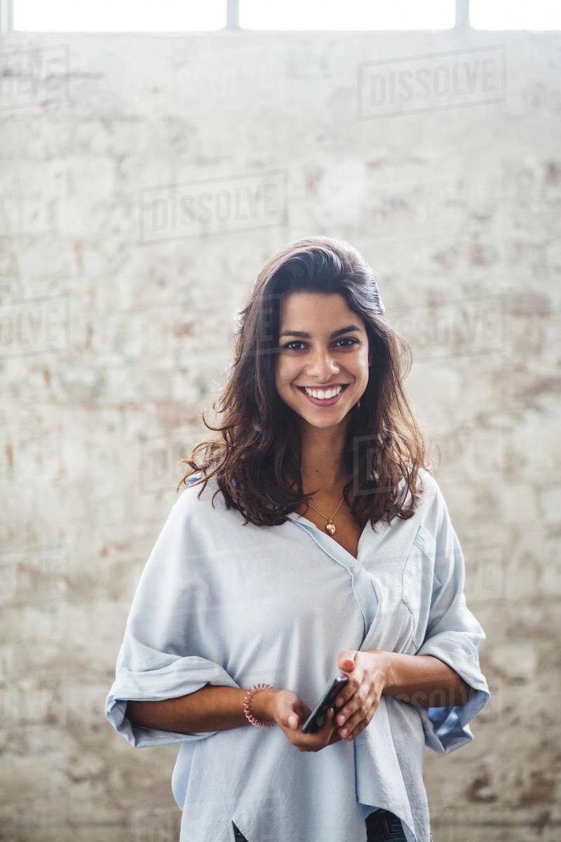 Portrait of smiling young female computer programmer holding smart phone standing against brick wall at creative office Royalty-free stock photo