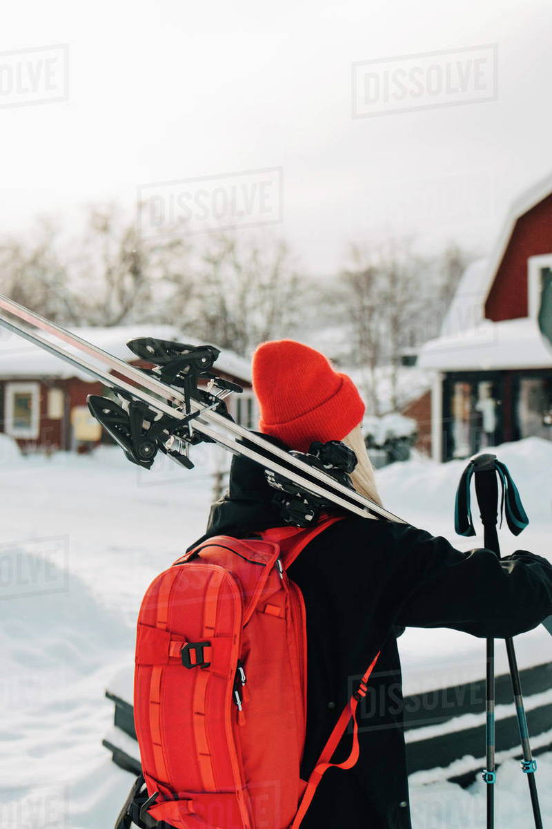 Rear view of young woman wearing red backpack carrying skis Stock