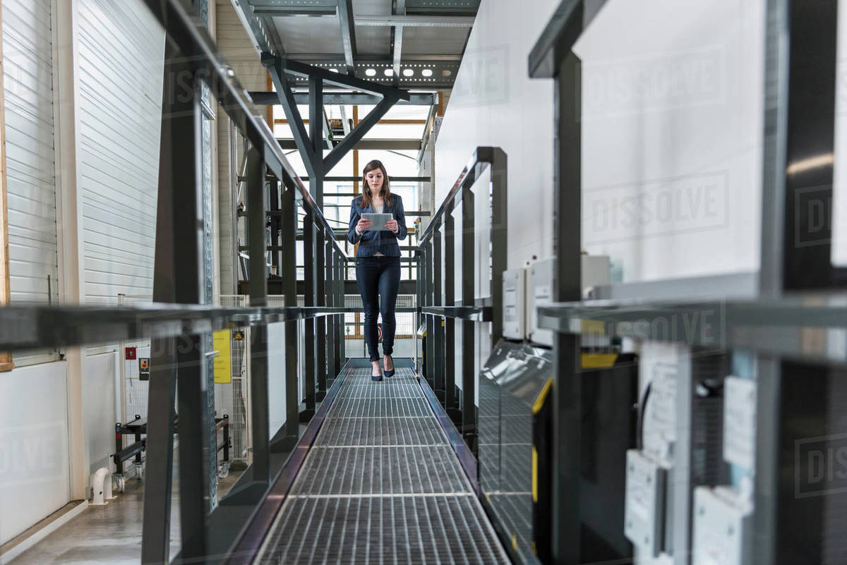 young woman walking along walkway in factory using digital tablet