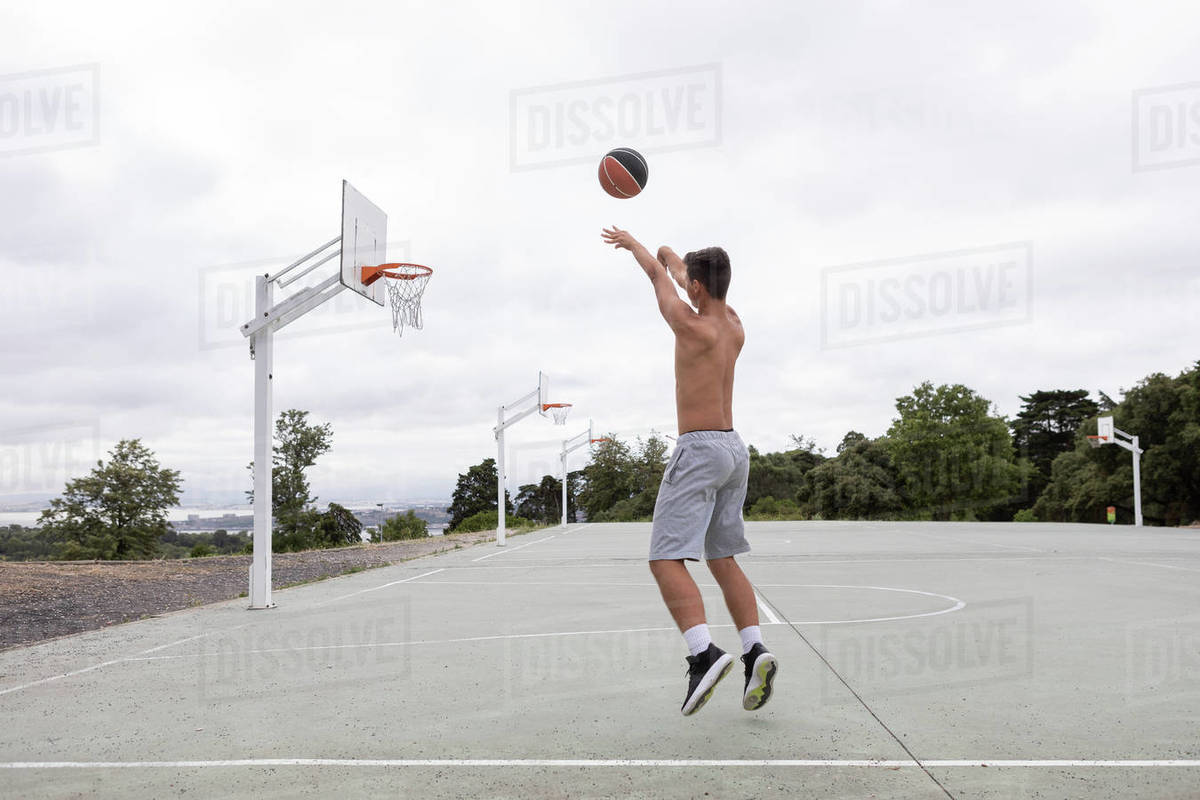 Male teenage basketball player jumping and throwing ball toward basketball hoop Stock Photo