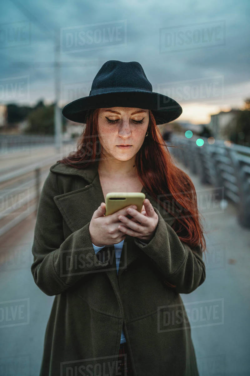 Young woman with long red hair on footbridge looking at smartphone at dusk Royalty-free stock photo