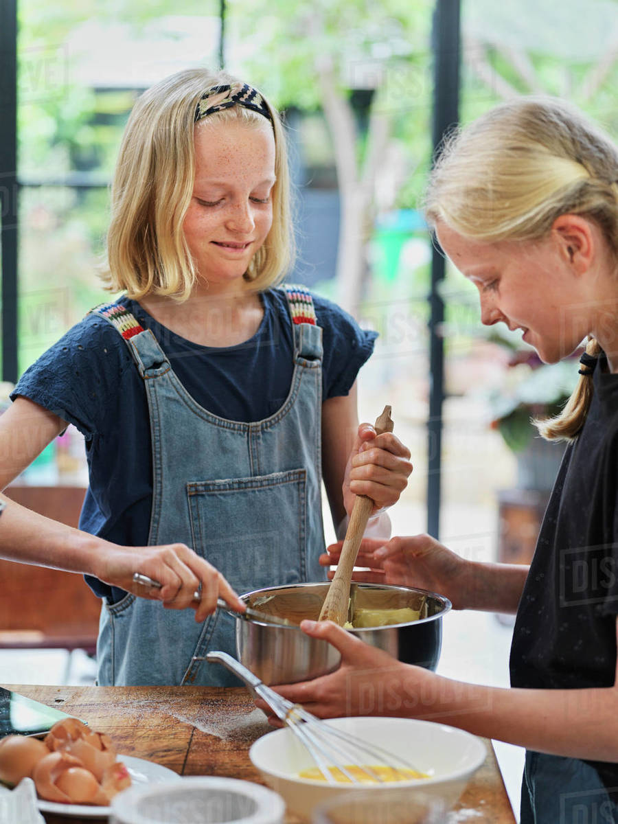 Girl and her sister baking a cake, stirring cake mixture in mixing bowl at kitchen table Royalty-free stock photo