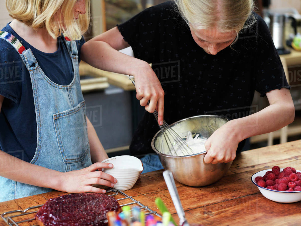 Girl and her sister baking a cake, whisking cream in mixing bowl at kitchen table Royalty-free stock photo